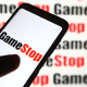 A cellphone with "GameStop" on the screen, held in front of a GameStop background.